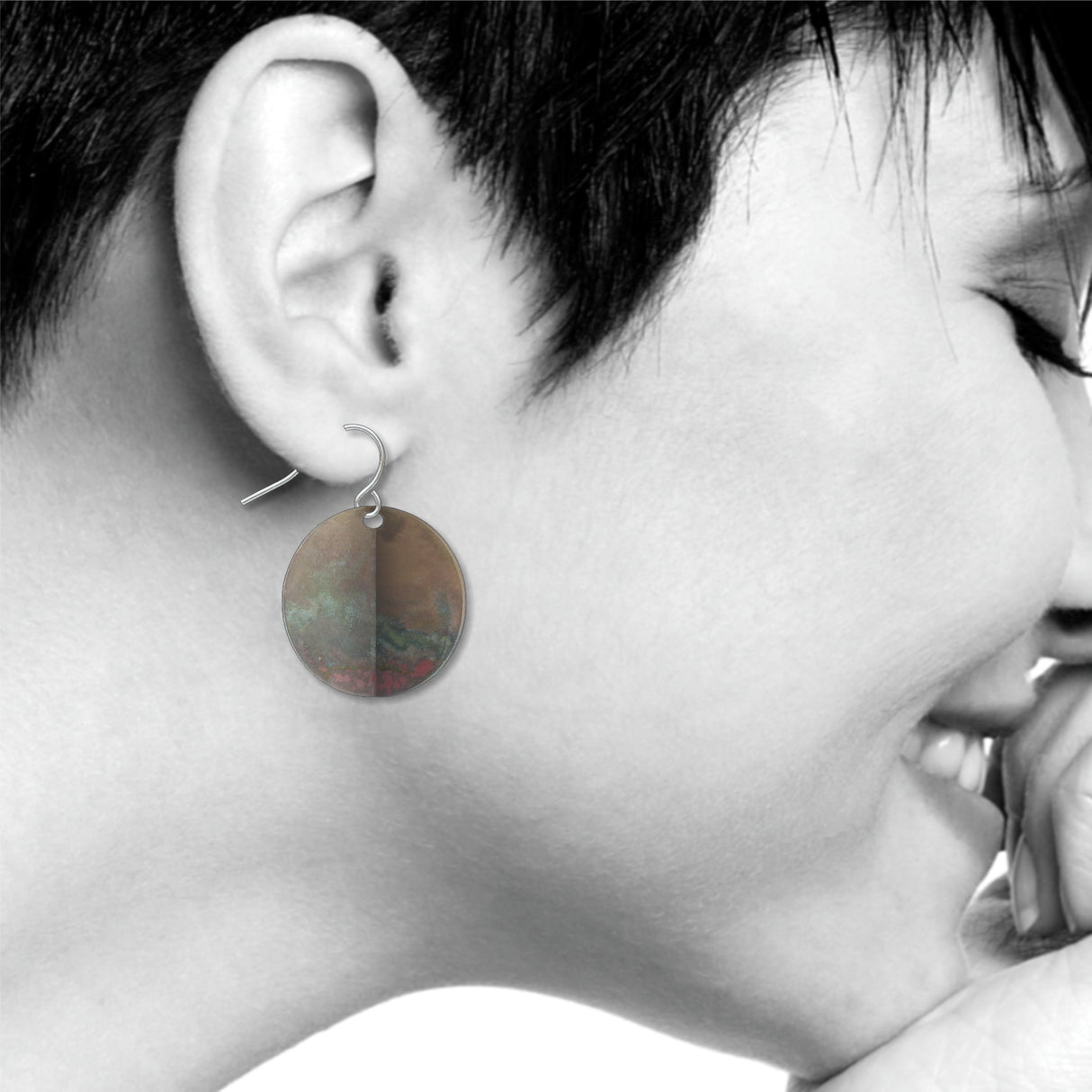 An Orb circular drop earring with a distressed metallic patina finish, shown worn on ear with an abstract modern art-inspired design