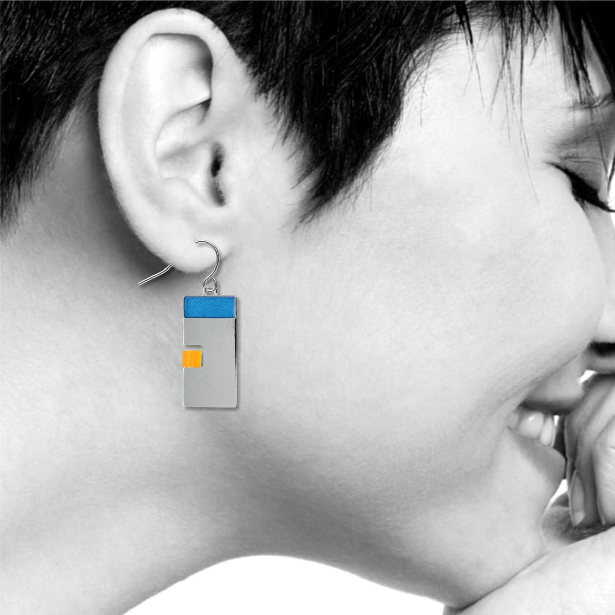 Model wearing a Poise geometric drop earring, featuring a sleek silver rectangle with a blue top bar and a small yellow accent, shown in a minimal, modern style.