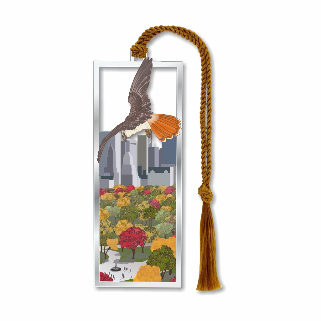 A silver metal bookmark featuring a design with a red-tailed hawk above Central Park, complete with a autumnal display of trees and a cityscape in the background. It includes a gold-colored tassel.