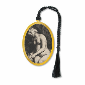 A metal bookmark featuring an etching of a nude woman by Rembrandt, surrounded by a gold oval border, with a black decorative tassel.